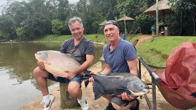 Dois amigos sentados na margem, cada um com seu peixe