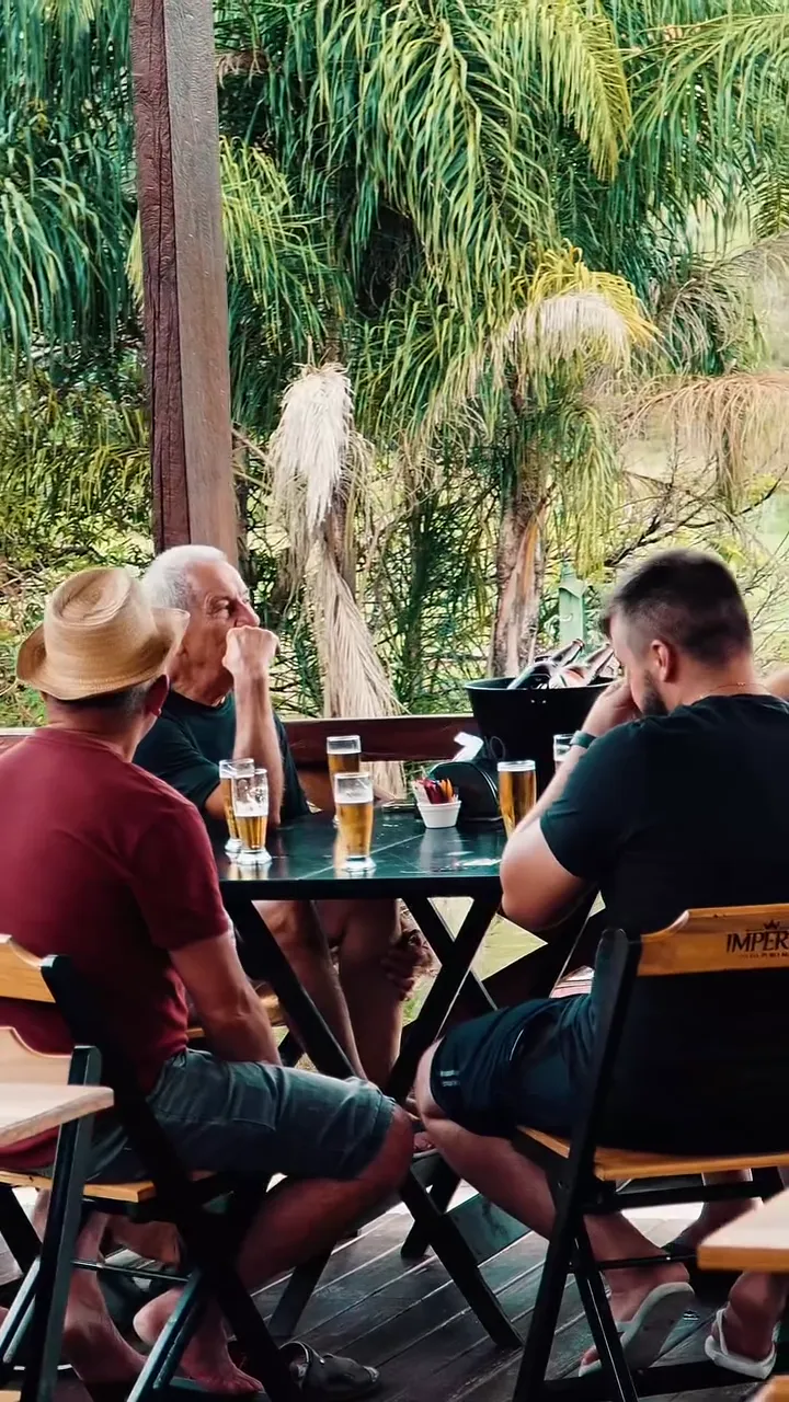 Amigos à mesa no deck, com bebidas e palmeiras ao fundo